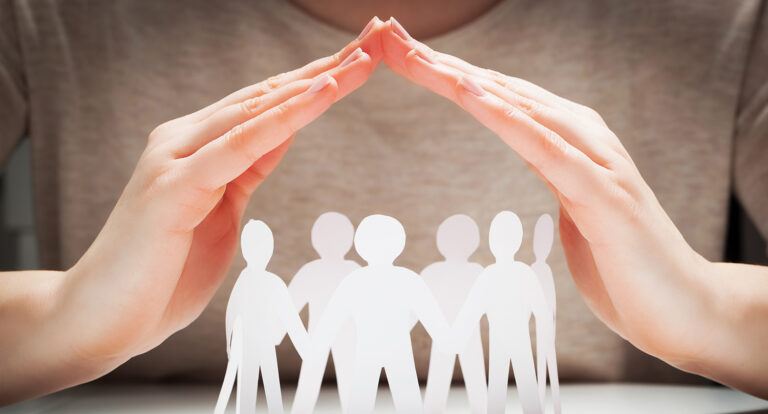 Ausgeschnittene Männchen halten sich an den Händen und werden von Händen überdacht. Symbol für Teamresilienz